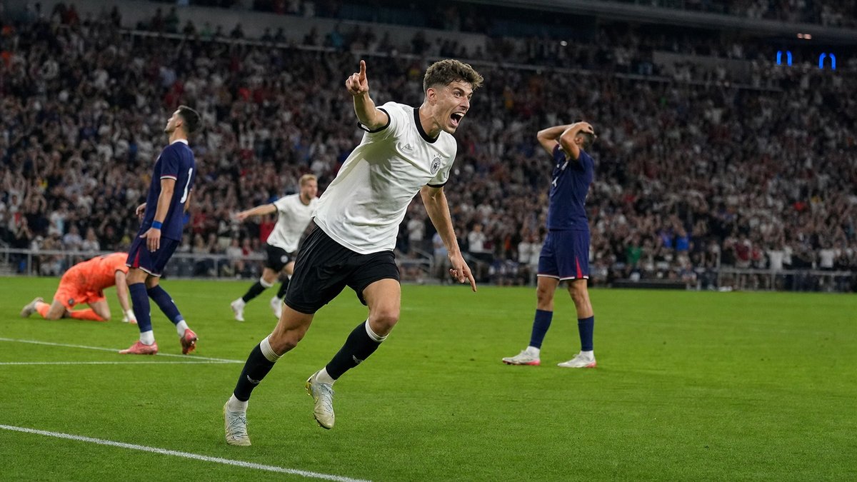 Jogador de futebol com uniforme branco comemora gol em estádio lotado enquanto adversários de azul lamentam.