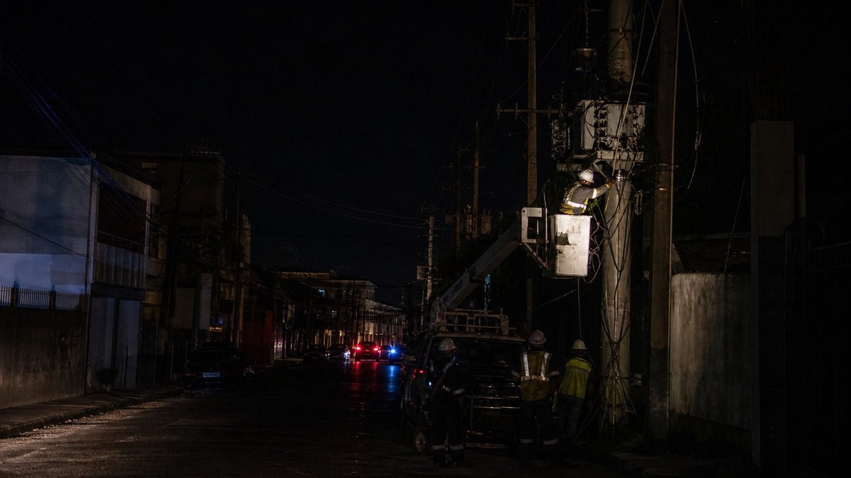 Rua urbana na Jamaica durante apagao noturno com equipes eletricas ao fundo