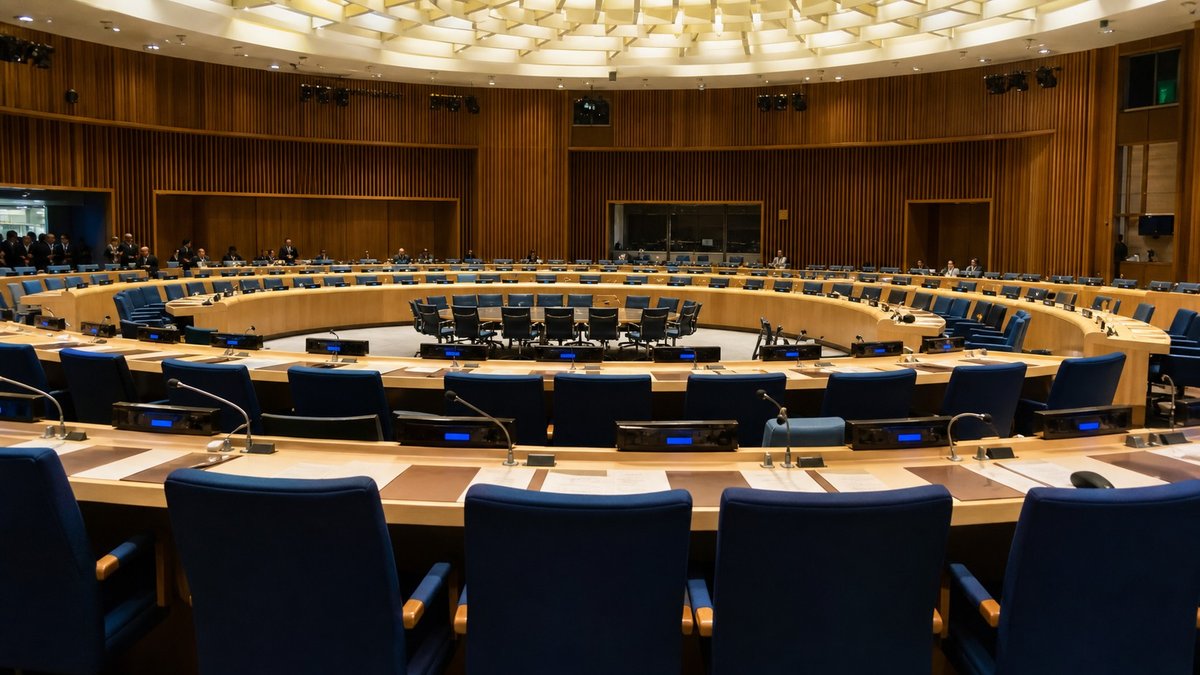 Plenário diplomático com mesas, microfones e cadeiras em uma sala de conferência internacional.