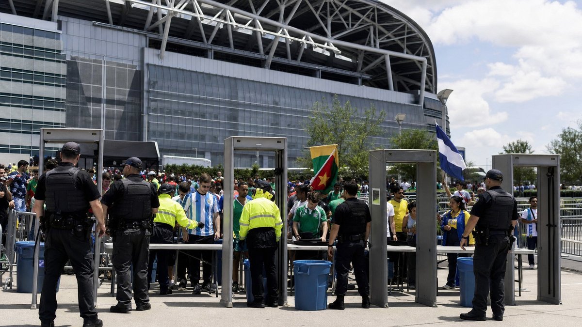 Seguranças e policiais organizam barreira de entrada diante de estádio moderno antes de jogo internacional.