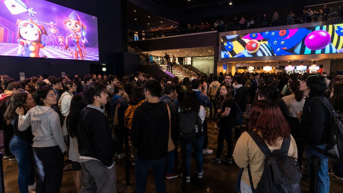 Fila de jovens em saguão de cinema com telas coloridas de animação digital ao fundo.