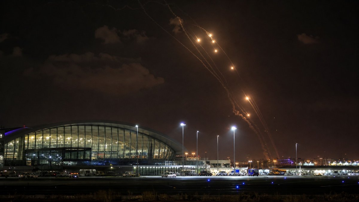 Terminal de aeroporto no Golfo à noite com luzes de emergência e interceptações distantes no céu.