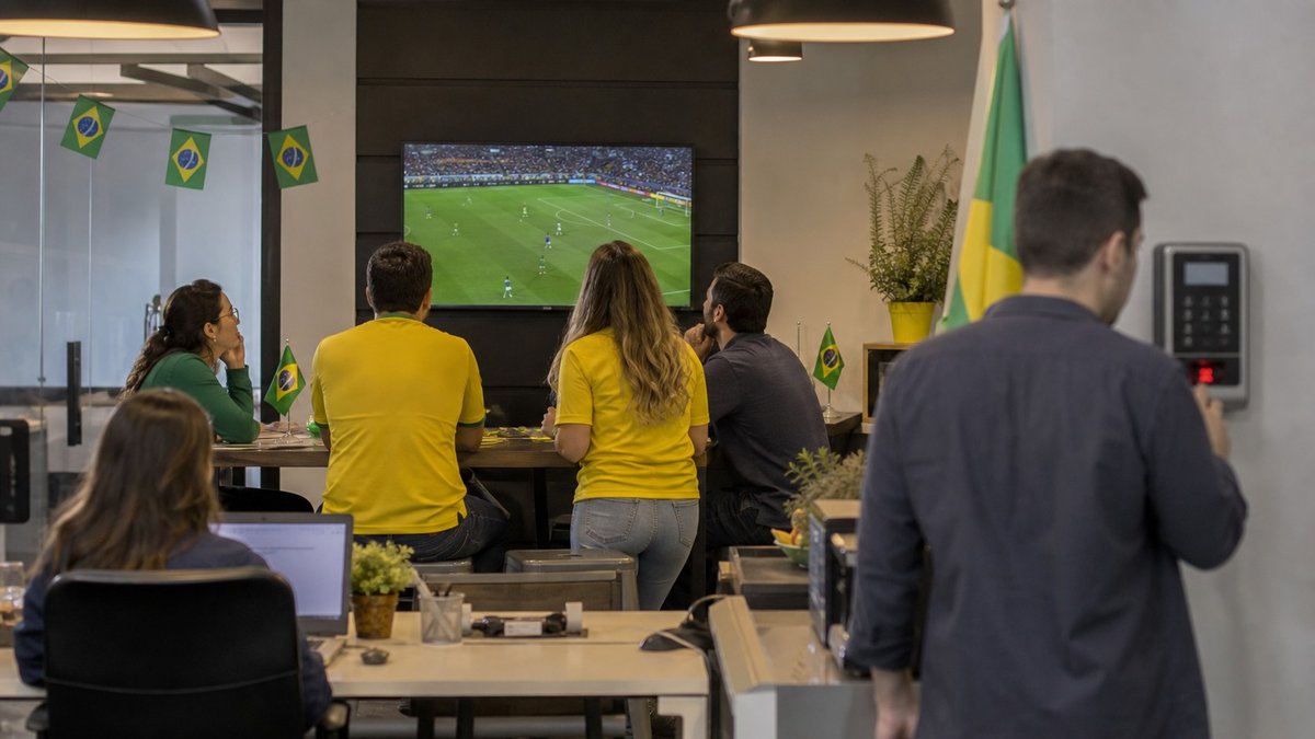 Torcedores brasileiros assistindo a jogo da Copa em ambiente de trabalho com bandeiras discretas.