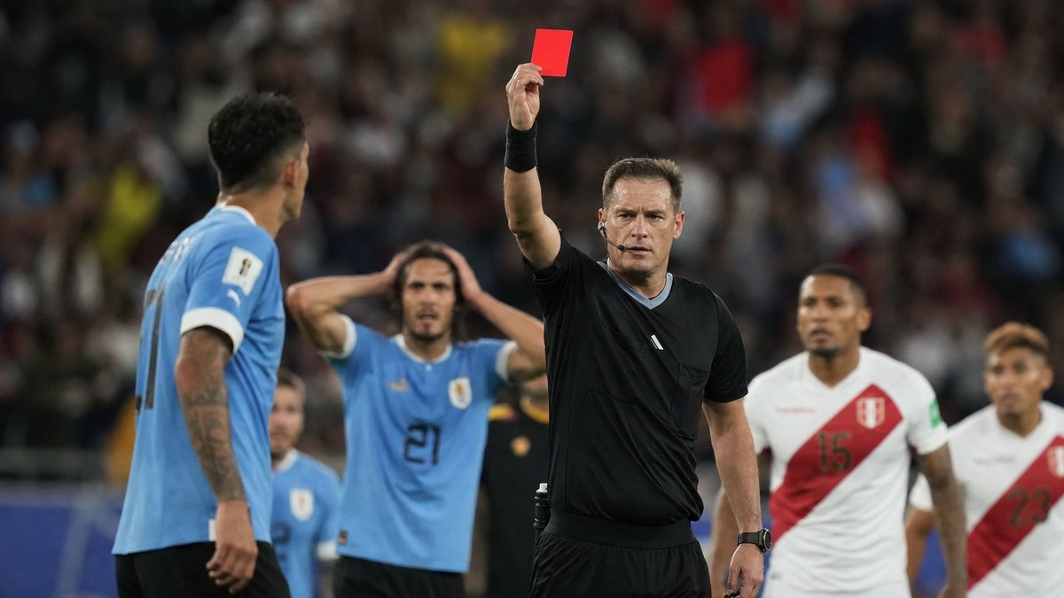 Árbitro de futebol erguendo cartão vermelho em estádio lotado durante jogo internacional à noite.